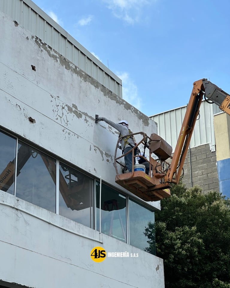 Construction worker in cherry picker lift cleaning weathered exterior facade of industrial building under clear sky